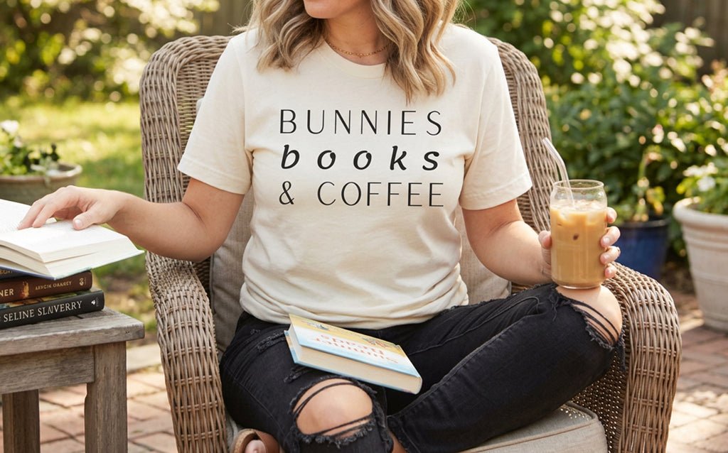 Curvy middle-aged woman with a blonde wavy bob, wearing a cream Bunnies, Books & Coffee t-shirt, relaxing on a patio chair with iced coffee and books.